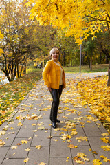 Fototapeta premium woman with short blonde hair stands in a park surrounded by autumn leaves. She wears a bright yellow jacket and black pants.