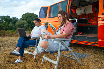 Women relaxing on chairs working with laptop and phone outside camper van.