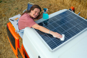 Happy woman cleaning solar panels on a camper van roof with a spray bottle and cloth.