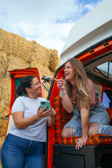 Two women looking at phone beside camper van and hay bales.