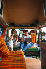 : Two women relaxing inside a camper van, sitting on orange plaid seats, talking and working on a laptop.