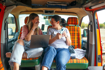 : Two women relaxing inside a camper van, sitting on orange plaid seats, talking and working on a laptop.