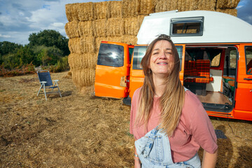 Woman smiling outdoors in front of orange camper van and hay bales.