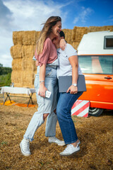 Two women laughing and hugging outdoors beside an orange camper van and hay bales.
