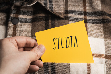 Yellow card with a handwritten inscription "Studia", a shaft in the hand, protruding from a brown plaid shirt (selective focus)