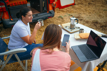 Two women drinking coffee and using smartphone and laptop at outdoor table.