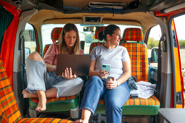Two women inside camper van working together using laptop and smartphone.