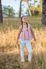 Young girl enjoying a sunny day in a field surrounded by trees and tall grass while wearing a striped top and white pants