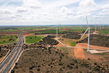 Wide aerial view of wind turbines on open fields next to highway. Renewable energy infrastructure integrated with rural landscape and transportation network