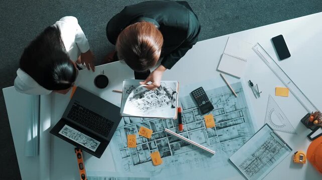 Top view of skilled project manager holding project plan and explain house design to architect engineer at meeting table with equipment, ruler, safety helmet and laptop scatter around. Alimentation.