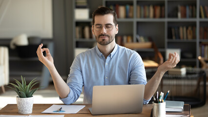 Calm young businessman meditating at workplace desk with closed eyes