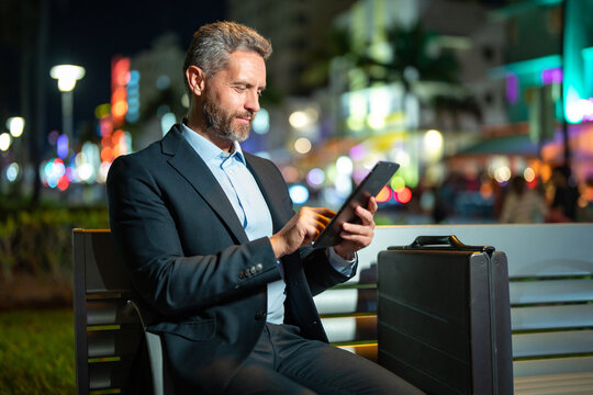 Online night business. Businessman working on tablet outdoors. Businessman with tablet outdoor. Businessman with a tablet on night street. Businessman using tablet, sit on bench. Night business.