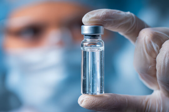 Medical professional wearing protective gear holds a transparent vial containing clear liquid for laboratory testing or vaccine preparation in clinical research setting