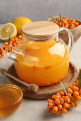 Sea buckthorn tea in teapot, fresh berries, honey and lemon on light grey table, closeup