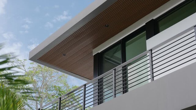 Top part of modern building. Flat roof with wooden slats underneath. Large glass windows with black frames and metal railing along balcony