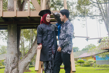 Indonesian couple in a happy moment and on a ladder from a tree house, with the man wearing typical Indonesian batik and the woman wearing an academic dress and decorated with a red hijab