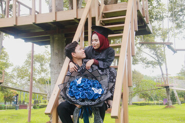 Indonesian couple in a happy moment and on a ladder from a tree house, with the man wearing typical Indonesian batik and the woman wearing an academic dress and decorated with a red hijab