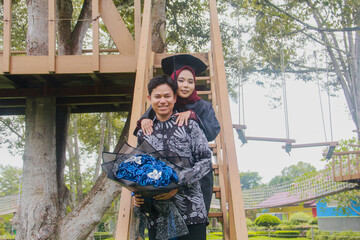 Indonesian couple in a happy moment and on a ladder from a tree house, with the man wearing typical Indonesian batik and the woman wearing an academic dress and decorated with a red hijab