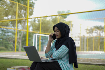 A young woman in a hijab uses her phone and laptop outdoors in a park, appearing focused on her work or studies amidst a natural setting