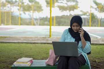 A young woman in a hijab uses her phone and laptop outdoors in a park, appearing focused on her work or studies amidst a natural setting