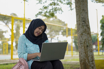 A young woman in a hijab uses her phone and laptop outdoors in a park, appearing focused on her work or studies amidst a natural setting