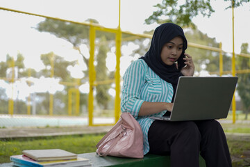 A young woman in a hijab uses her phone and laptop outdoors in a park, appearing focused on her work or studies amidst a natural setting