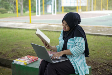 A young woman in a hijab uses her phone and laptop outdoors in a park, appearing focused on her work or studies amidst a natural setting
