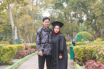 Indonesian couple in a happy moment in a park with the man wearing traditional Indonesian batik and the woman wearing an academic dress and decorated with a red hijab