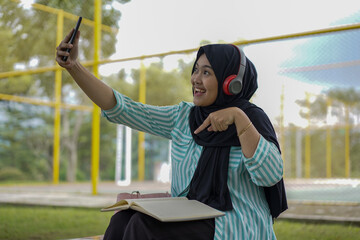 Young woman in hijab and headphones using a smartphone to record a video outdoors with a notebook and pen nearby, capturing a moment of content creation and learning in a casual setting
