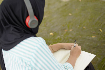A person wearing headphones and writing in a notebook near a flowing body of water, possibly a stream or river, engaged in a creative pursuit