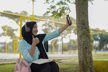 Young woman in hijab and headphones using a smartphone to record a video outdoors with a notebook and pen nearby, capturing a moment of content creation and learning in a casual setting