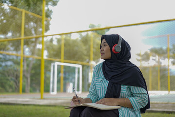 Serene young woman in a hijab and red headphones, deeply engrossed in reading a book while studying outdoors on a tranquil park bench, fostering knowledge and personal growth