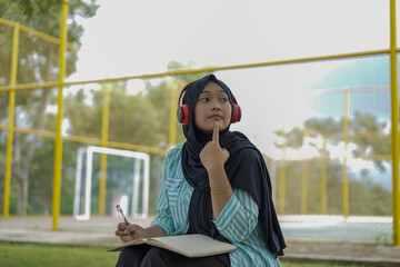 Serene young woman in a hijab and red headphones, deeply engrossed in reading a book while studying outdoors on a tranquil park bench, fostering knowledge and personal growth