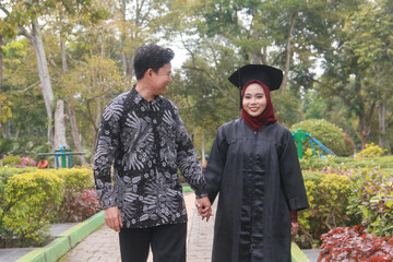 Indonesian couple in a happy moment in a park with the man wearing traditional Indonesian batik and the woman wearing an academic dress and decorated with a red hijab