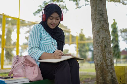 Serene young woman in a hijab and red headphones, deeply engrossed in reading a book while studying outdoors on a tranquil park bench, fostering knowledge and personal growth