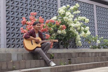 Outdoor shot of attractive Asian man playing guitar at city park in the morning