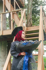Beautiful Indonesian Muslim woman wearing maroon hijab and toga and academy dress appears elegant in a park
