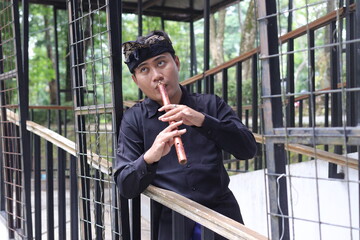 Outdoor shot of Asian man playing traditional bamboo flute in city park with green surroundings