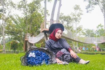 Beautiful Indonesian Muslim woman wearing maroon hijab and toga and academy dress sitting on the grass and accompanied by a blue bouquet of flowers