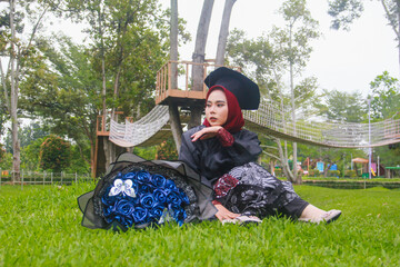 Beautiful Indonesian Muslim woman wearing maroon hijab and toga and academy dress sitting on the grass and accompanied by a blue bouquet of flowers