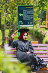 Beautiful Indonesian Muslim woman wearing maroon hijab and toga and academy dress sitting on a bench in a park