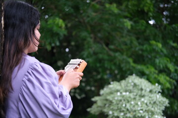 Asian woman relaxes in a city park on a sunny summer day, playing a traditional kalimba. Surrounded by greenery, she enjoys a peaceful moment, blending music with the tranquility of nature