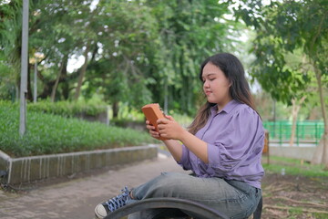 Asian woman relaxes in a city park on a sunny summer day, playing a traditional kalimba. Surrounded by greenery, she enjoys a peaceful moment, blending music with the tranquility of nature