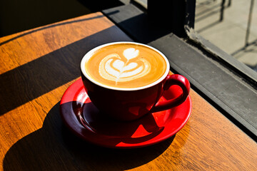Close up view of hot cafe latte on the wooden table. red cup or mug. art. brown. dark, black, shadows. morning. relax, coffee time. cafe. coffee shop. sun shine, light. ceramic.