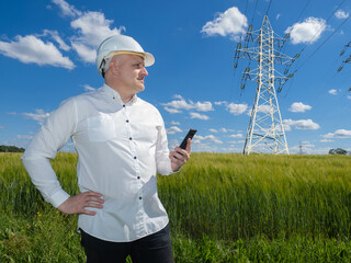 Man engineer stands near high-voltage power line. Worker constructs energy transmission infrastructure. Businessman in helmet in green field. Man engineer with phone inspects power line support.