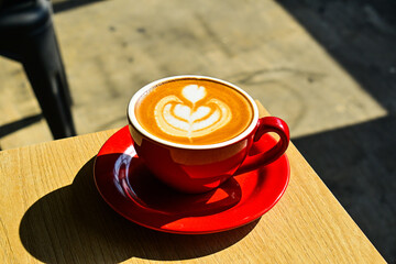 Close up view of hot cafe latte on the cornerwooden table. red cup or mug. art. brown. dark, black, shadows. morning. relax, coffee time. cafe. coffee shop. sun shine, light. ceramic.