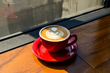 Close up view of hot cafe latte on the wooden table. red cup or mug. art. brown. dark, black, shadows. morning. relax, coffee time. cafe. coffee shop. sun shine, light. ceramic.