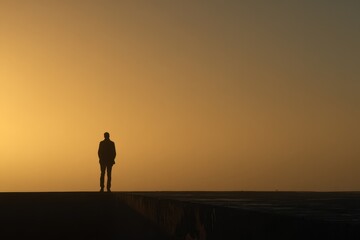 Fototapeta premium Silhouette of man standing alone on elevated path against golden sunset sky, expressing solitude, contemplation, and peaceful reflection in minimalist evening landscape
