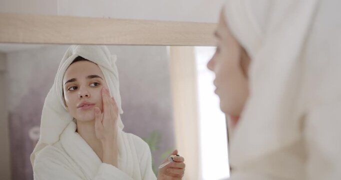 Young beautiful woman in a bathrobe and a towel on head applying face lotion and skincare cream, serum, she prepares to be beautiful looking in the mirror at home. Woman applying rejuvenating serum.