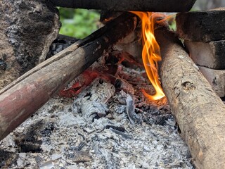 Close-up of a campfire with burning logs and glowing embers, surrounded by ash and rocks.